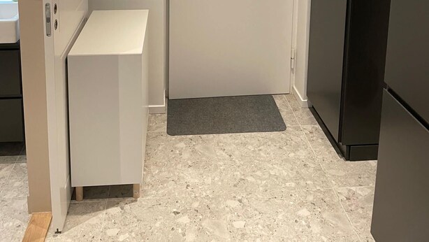 Une pièce de taille modeste avec une porte blanche, un réfrigérateur noir, un petit meuble blanc et un tapis gris sur le sol.