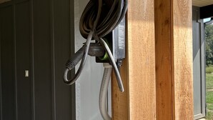 Un poste de madera con un cable enrollado y un dispositivo verde sujeto, probablemente una bomba de agua, en un espacio exterior cubierto con suelo de piedra y una pared gris al fondo.