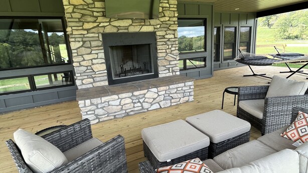 Un patio exterior con mobiliario de mimbre, una chimenea de piedra y grandes ventanales que ofrecen vistas al exterior.
