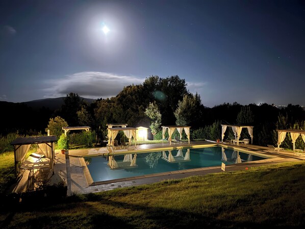 A swimming pool with a pergola and lounge chairs, surrounded by grass and trees, under a night sky with a visible moon.