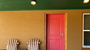 A red door with two wooden Adirondack chairs and a small table outside a building with a green roof and brick walls.