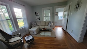 A living room with a sectional sofa, a coffee table, and a chair. There are two windows with curtains, a door, and a wall-mounted mirror.