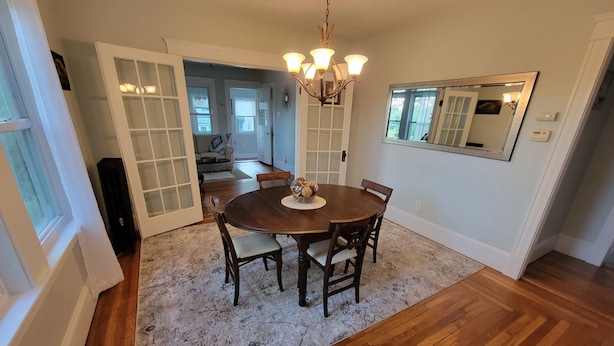 A dining area with a round wooden table, four chairs, a chandelier, and a large mirror on the wall.