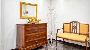 Een houten dressoir met lades, een spiegel met gouden lijst, een stoel met geel gestoffeerde bekleding en een staande lamp.