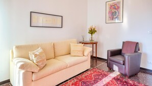 Een woonkamer met een beige bank, een donkerblauwe fauteuil, een rood geweven vloerkleed, een klein rond tafeltje met een bloevas, en twee ingelijste kunstwerken aan de muur.