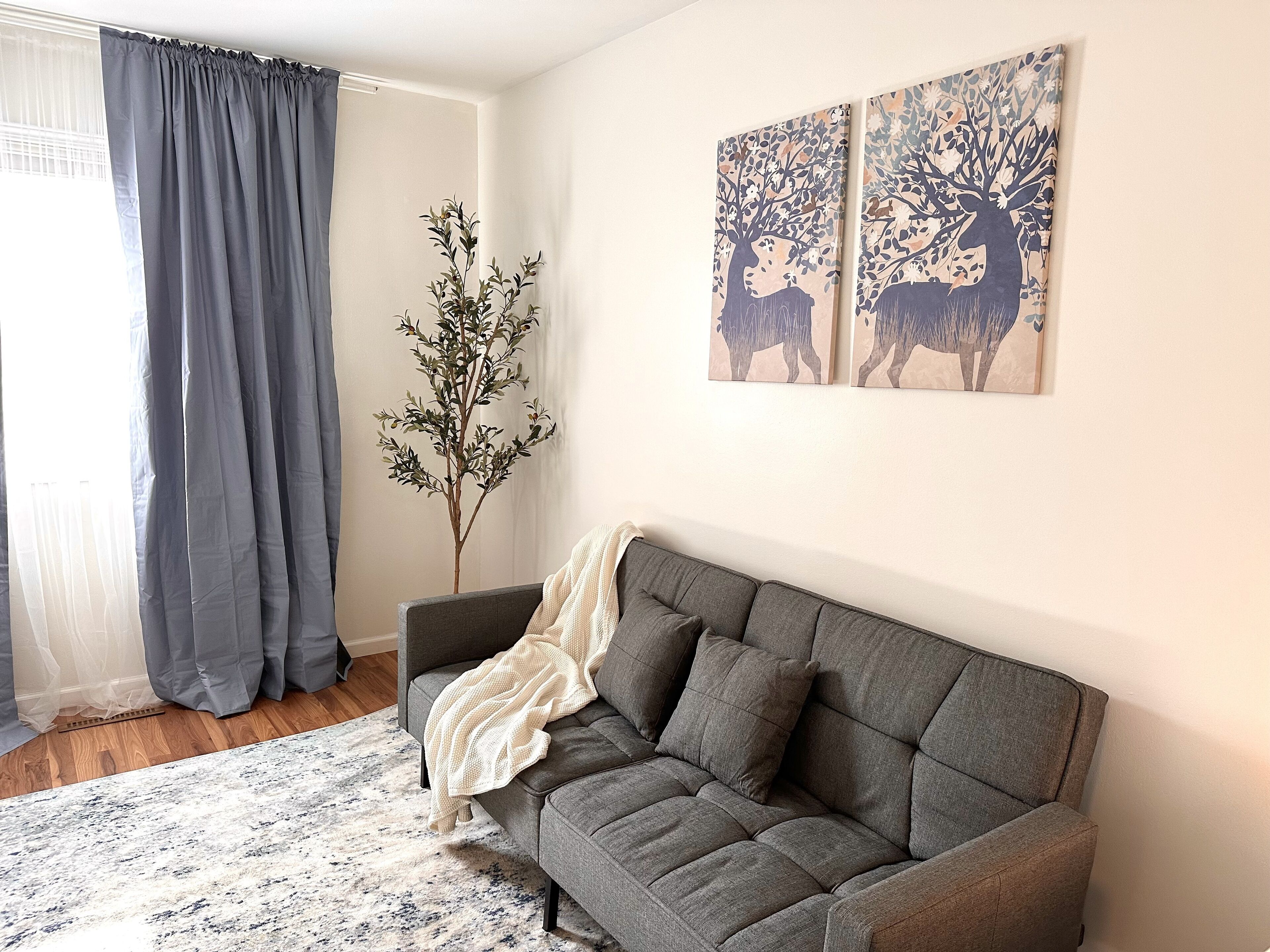 A living room with a grey sofa, a white rug, a plant, and two wall art pieces of deer.