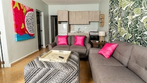 Un salon avec un canapé, un pouf et une table basse. Au fond, on aperçoit une cuisine avec des meubles en bois et une cuisinière.
