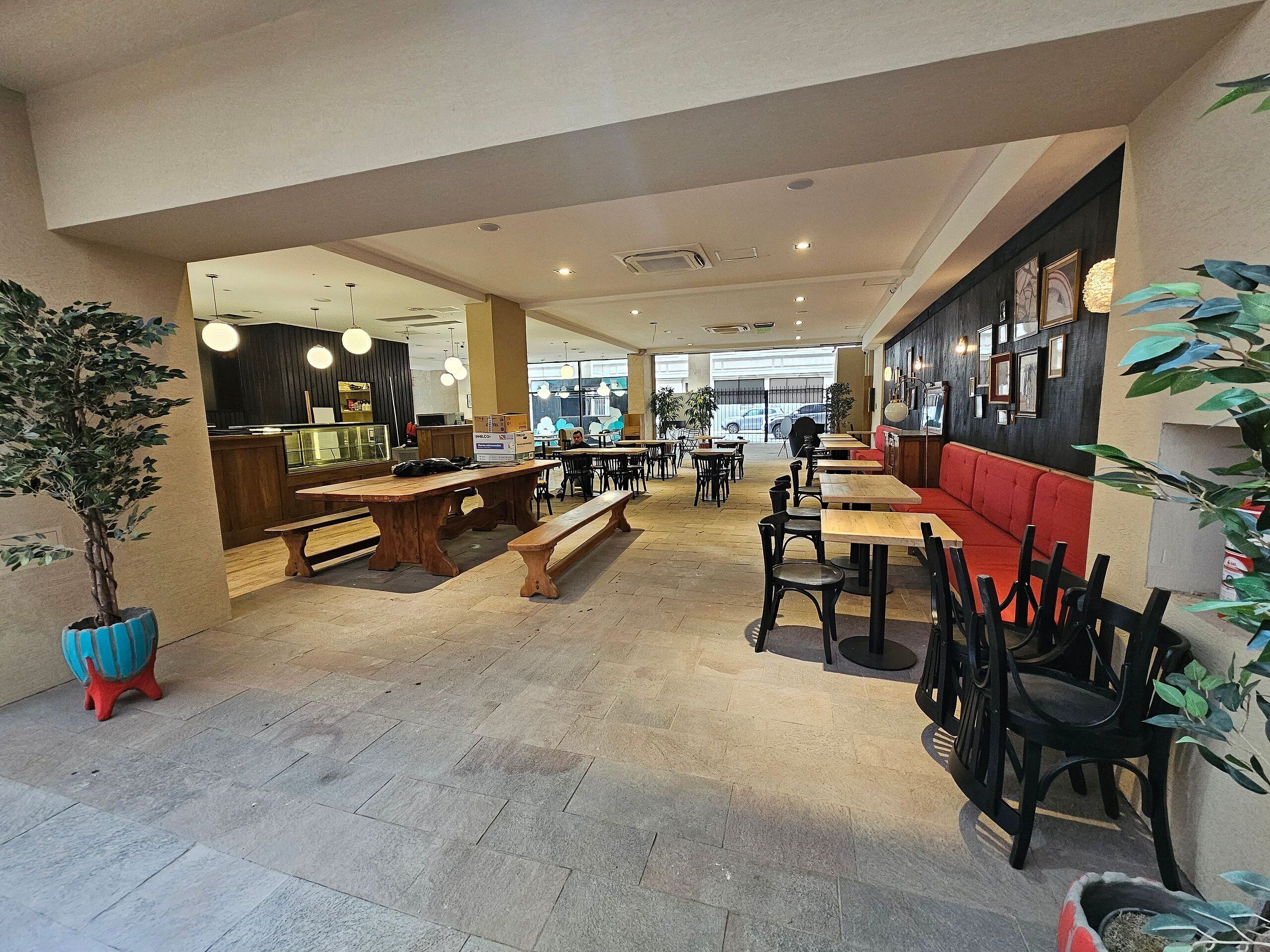 A spacious cafe with wooden tables and benches, a counter area, and a seating area with red cushions.
