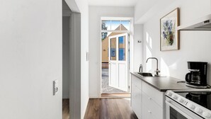 A modern kitchen with a built-in oven, a coffee maker, and a view through an open door to an outdoor area. - Lovely apartment at Solgaarden (9) Årsdale (Svaneke)