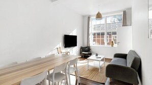 A modern living space with a wooden dining table, white chairs, a grey sofa, a small white table, and a TV mounted on the wall. - Lovely apartment at Solgaarden (9) Årsdale (Svaneke)