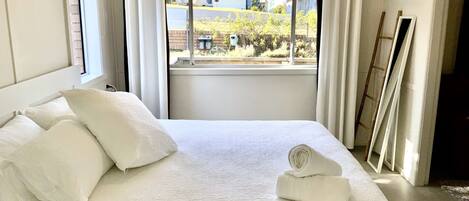 A hotel room with a large bed, white linens, a view of the outside, and a ladder leaning against the wall.