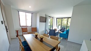 A modern dining area with a wooden table and chairs, a television mounted on the wall, and a view of the outdoors through large windows.