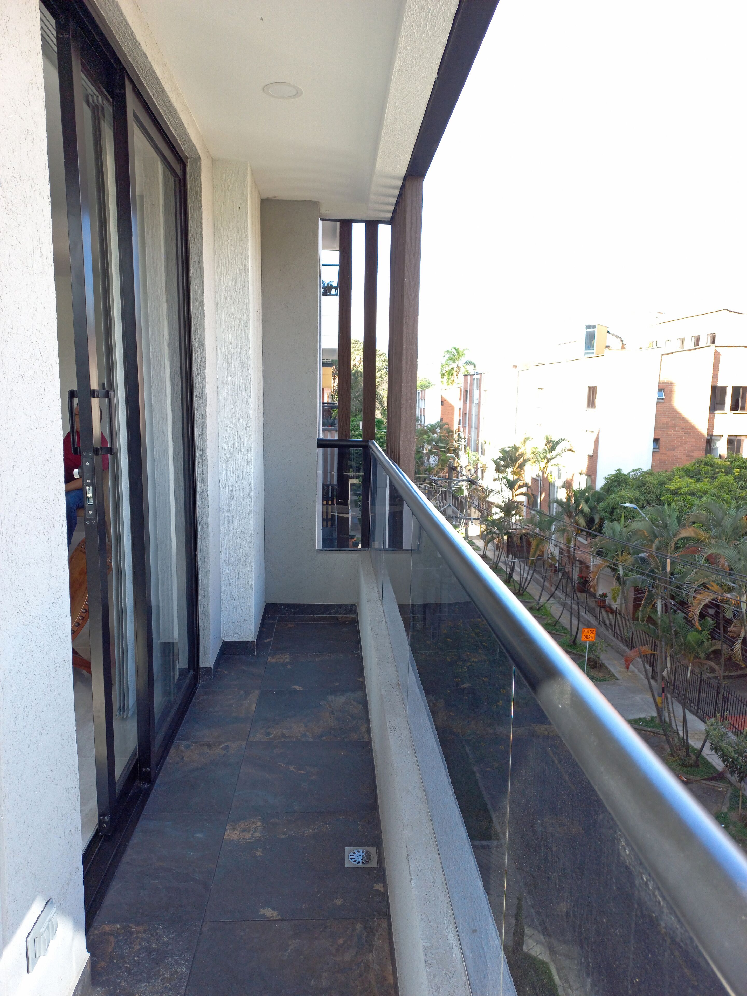 A narrow balcony with a glass door leading to a room, a metal railing, and a view of buildings and greenery.