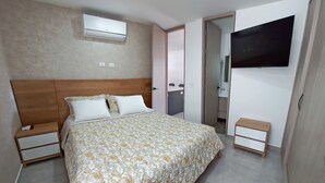 A bedroom with a bed, bedside tables, an air conditioning unit, a television, and a bathroom visible through an open door.