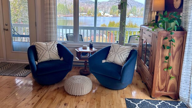Un salón acogedor con dos sillones azules, un armario de madera y vistas a un lago y montañas a través de amplios ventanales.