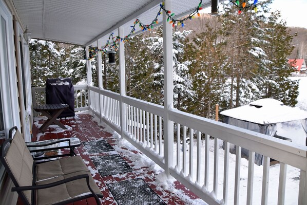 A covered porch with a bench, a table, and a snow-covered landscape with trees and a building in the background.