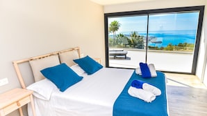 Ein modernes Hotelzimmer mit einem großen Bett, einem Nachttisch aus Holz und Blick auf das Meer durch raumhohe Fenster.