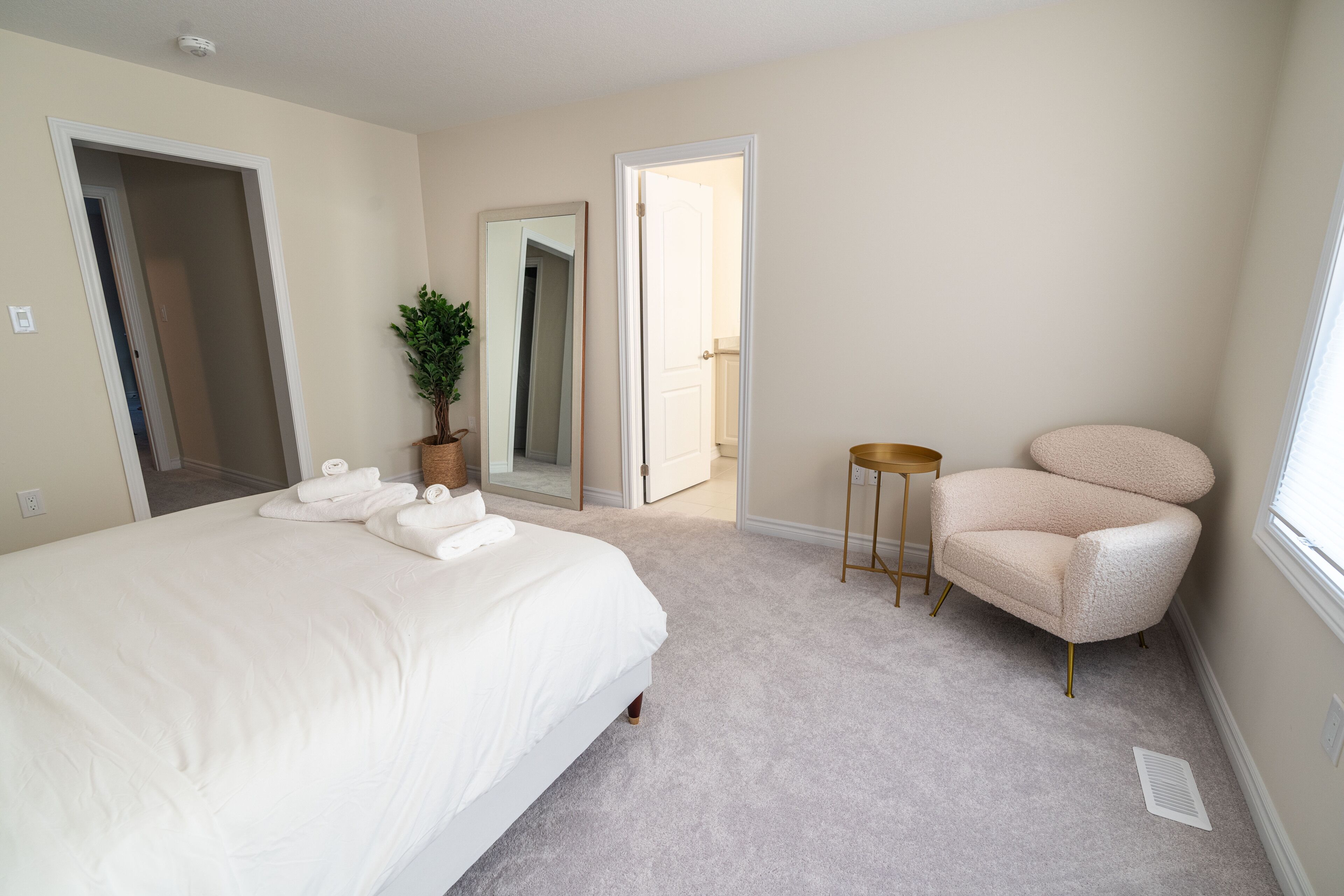 A bedroom with a large bed, a chair, a small table, a potted plant, and a mirror.