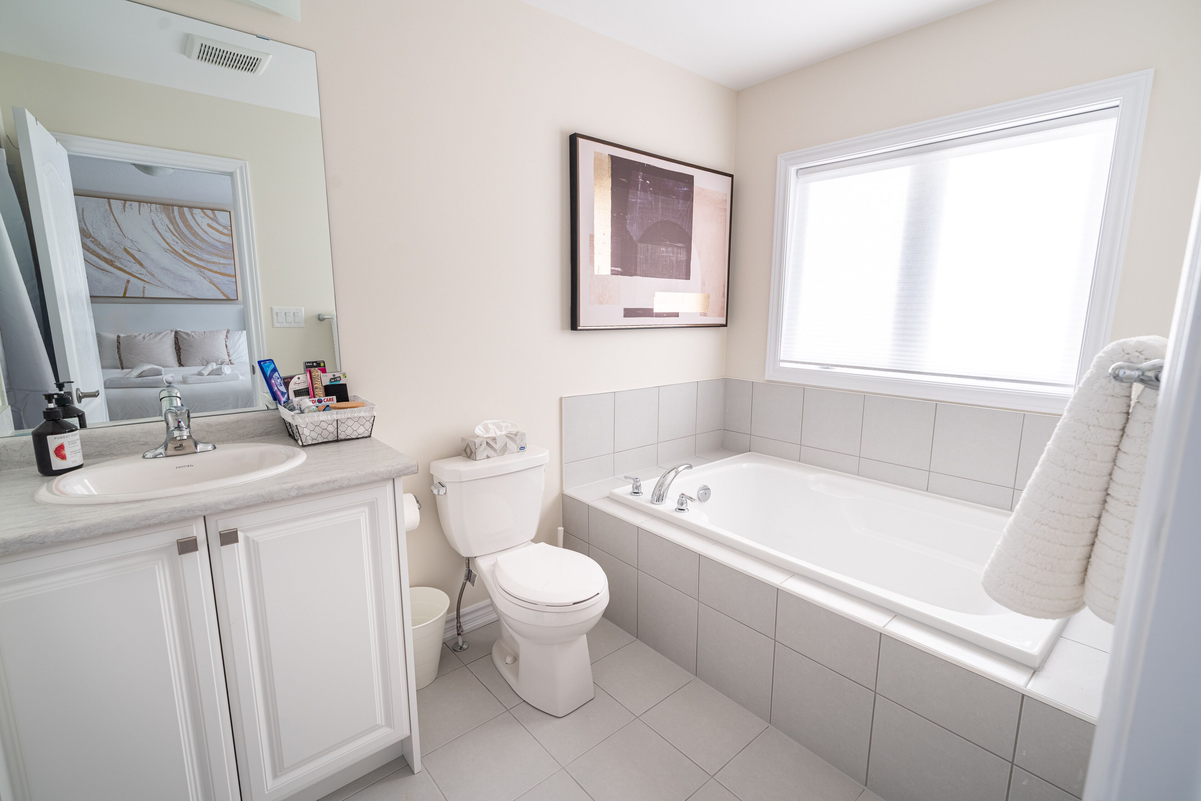 Un baño moderno con lavamanos, inodoro y bañera blancos. Hay un cuadro en la pared y una ventana que deja entrar luz natural.