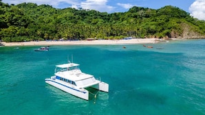 A white boat with a blue stripe in turquoise waters, surrounded by a beach and dense green forest. - Beachfront Luxury Condo with Direct Access to the Beach, 3 BR, 2 Baths, Balcony! (Jacó)