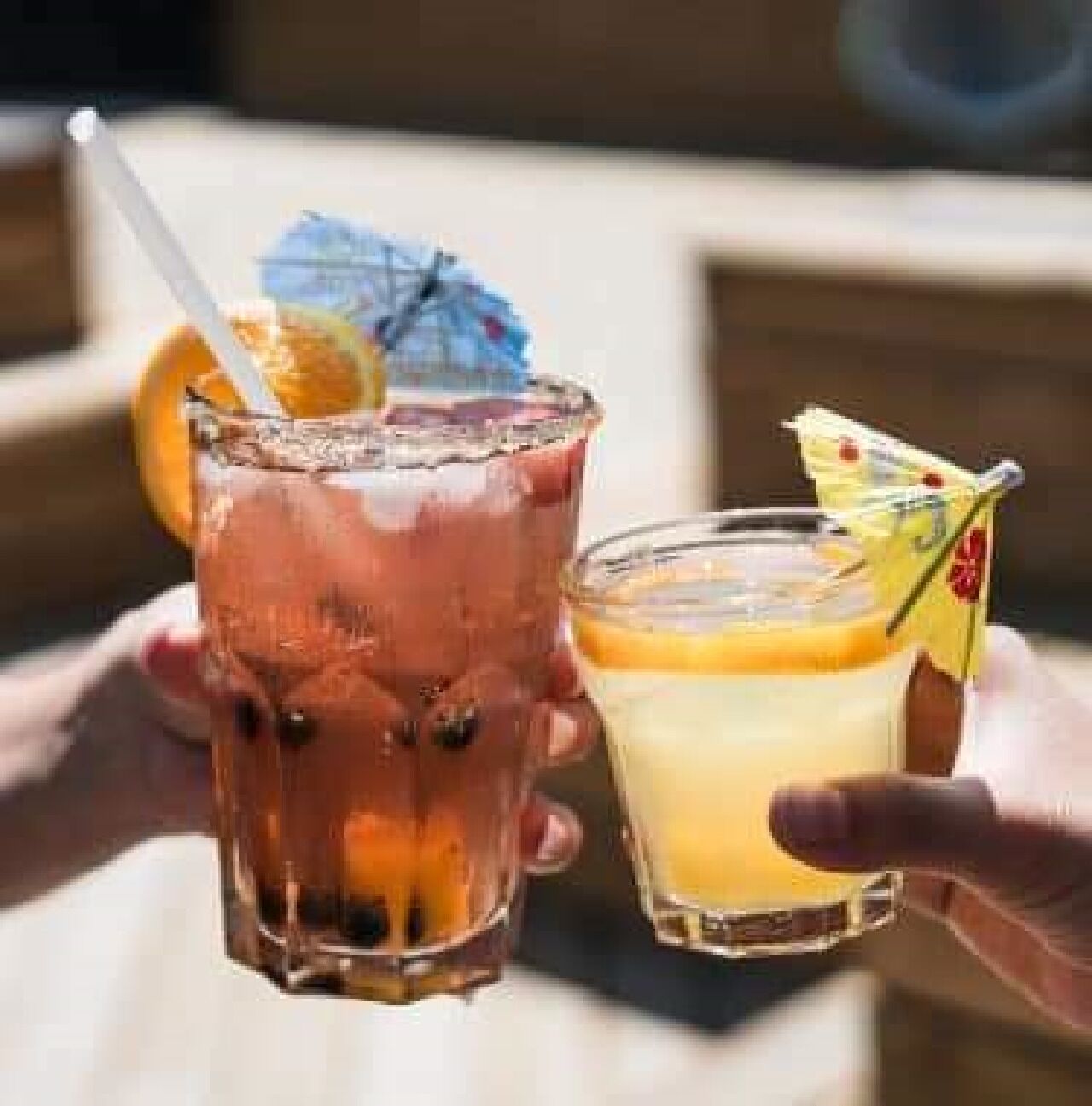 Two drinks, one in a tall glass and one in a short glass, with a straw and a slice of orange in the tall glass.