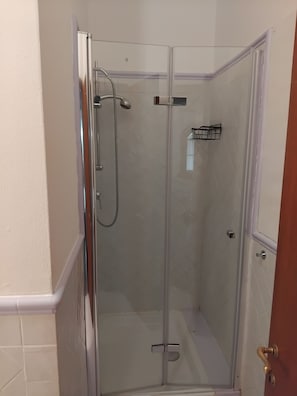 A glass-enclosed shower stall with a chrome showerhead and a handheld showerhead, a white tiled wall, and a door with a brass handle. - Apartment with Garden View (GAMBASSI TERME)