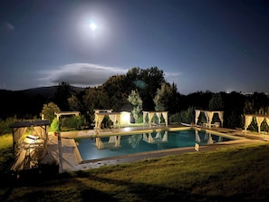 Une piscine agrémentée d’une pergola et de chaises longues, entourée de pelouse et d’arbres, sous un ciel nocturne où l’on peut apercevoir la lune.
