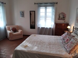 Un dormitorio con cama, una silla, cómoda, espejo y ventana con cortinas.