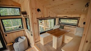 A wooden cabin interior with a dining table, seating area, and large windows offering views of the forest. - Luxury Treehouse central Stowe/Waterbury. Borders State park. Walk to the Lake! (Waterbury Center)