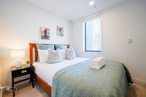 A bedroom with a bed, a nightstand, a lamp, a window, and two wall art pieces. - 2-Bs Suite Adjacent to Sky Tower (Auckland)