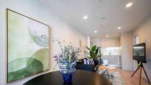 A modern living room with a round table, a large abstract painting, a vase with flowers, a sofa, and a kitchen in the background.