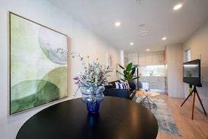 Ein modernes Wohnzimmer mit einem runden Tisch, einem großen abstrakten Gemälde, einer Vase mit Blumen, einer Couch und einer Küche im Hintergrund.