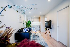 Un salon moderne avec un canapé noir, un téléviseur posé sur un pied, et une cuisine en arrière-plan.