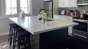 A modern kitchen with white cabinets, a large island with stools, a stainless steel refrigerator, and a built-in oven. - Sunrise net on Pigeon Lake 4 season cottage (Kawartha Lakes)