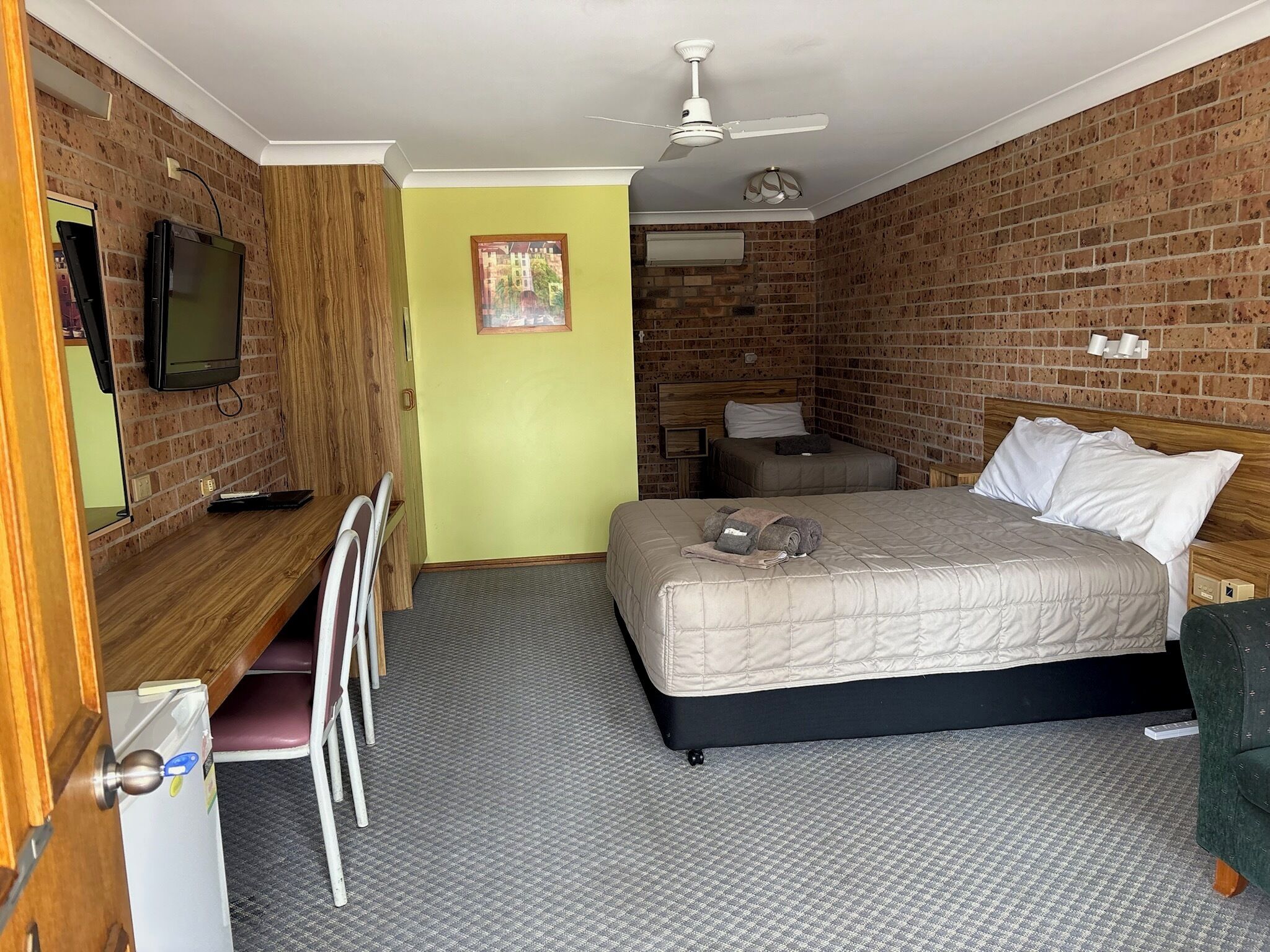 A hotel room with a brick wall, a bed with pillows, a desk with a chair, a television, and a ceiling fan.