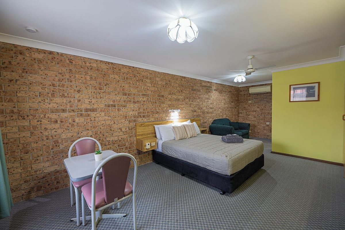 A bedroom with a brick wall, a bed, a desk with a chair, and a ceiling fan.