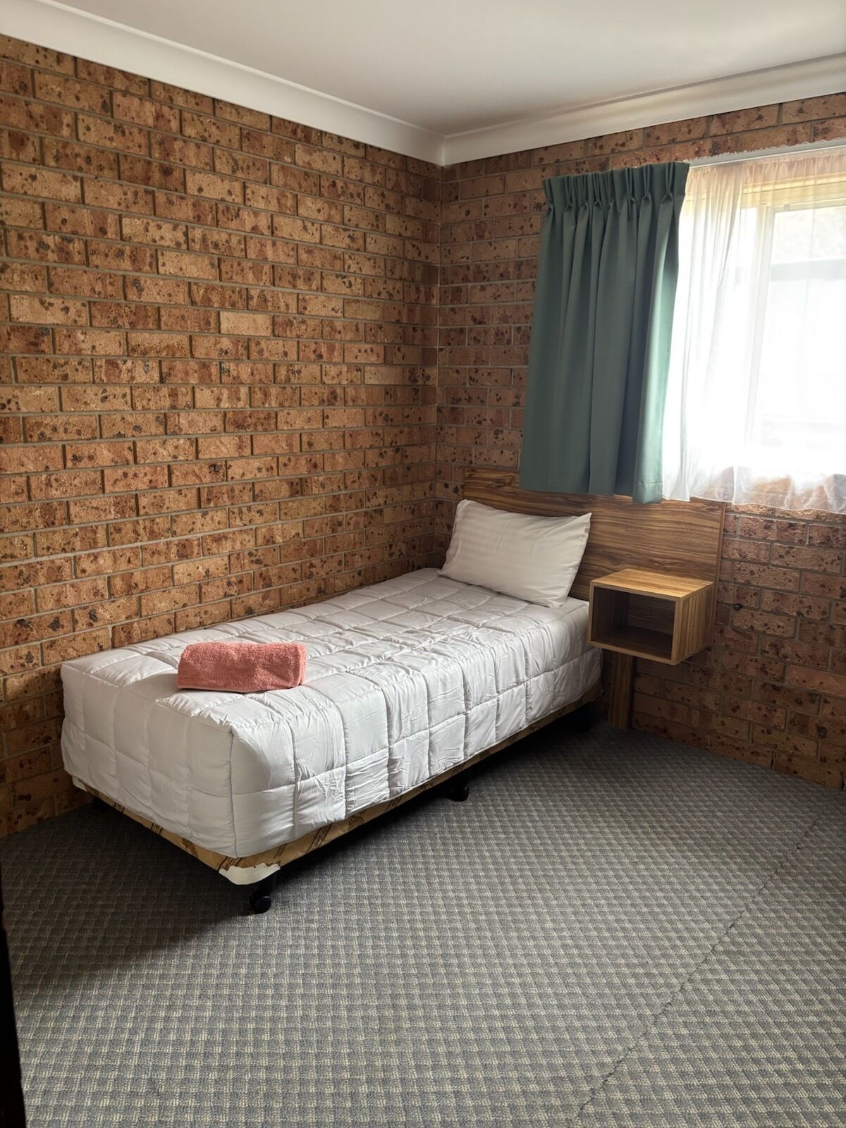 Ein kleines Zimmer mit einem Einzelbett, einer Ziegelwand, einem Holznachttisch und einem Fenster mit durchscheinendem Vorhang.