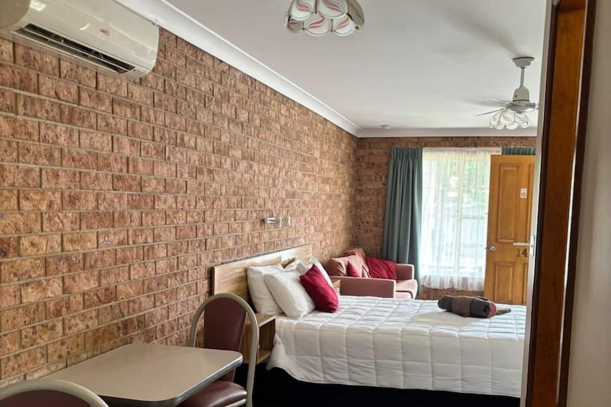 A room with a brick wall, a bed with red pillows, a desk with a chair, and a ceiling fan.