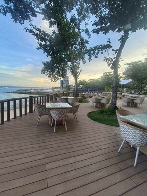 An outdoor deck with tables and chairs, overlooking a body of water and a distant cityscape. - One bedroom Unit at mactan newtown (Lapu-Lapu)