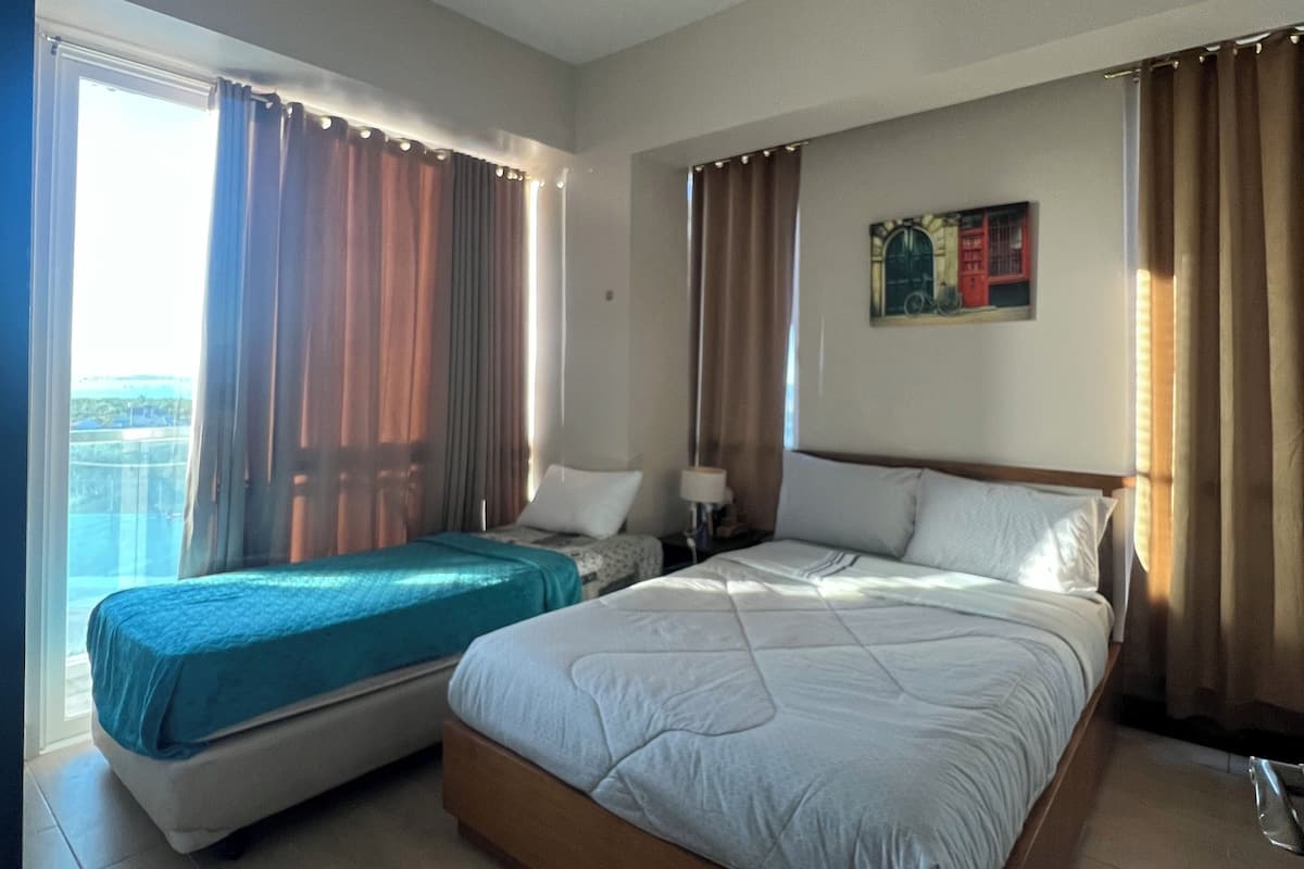 A hotel room with two beds, a large window with curtains, a painting on the wall, and a ceiling light.