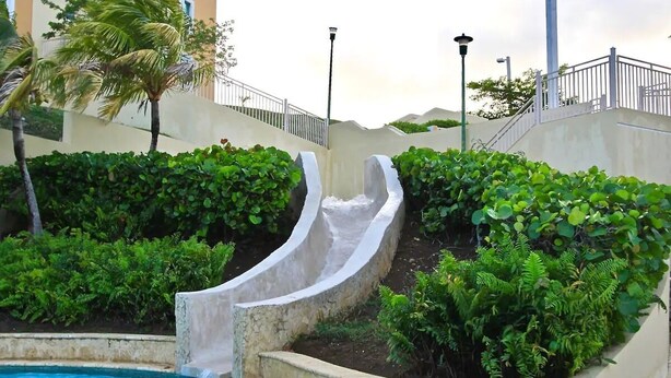 Une piscine avec un toboggan, entourée d’une végétation luxuriante, avec un bâtiment visible au loin.