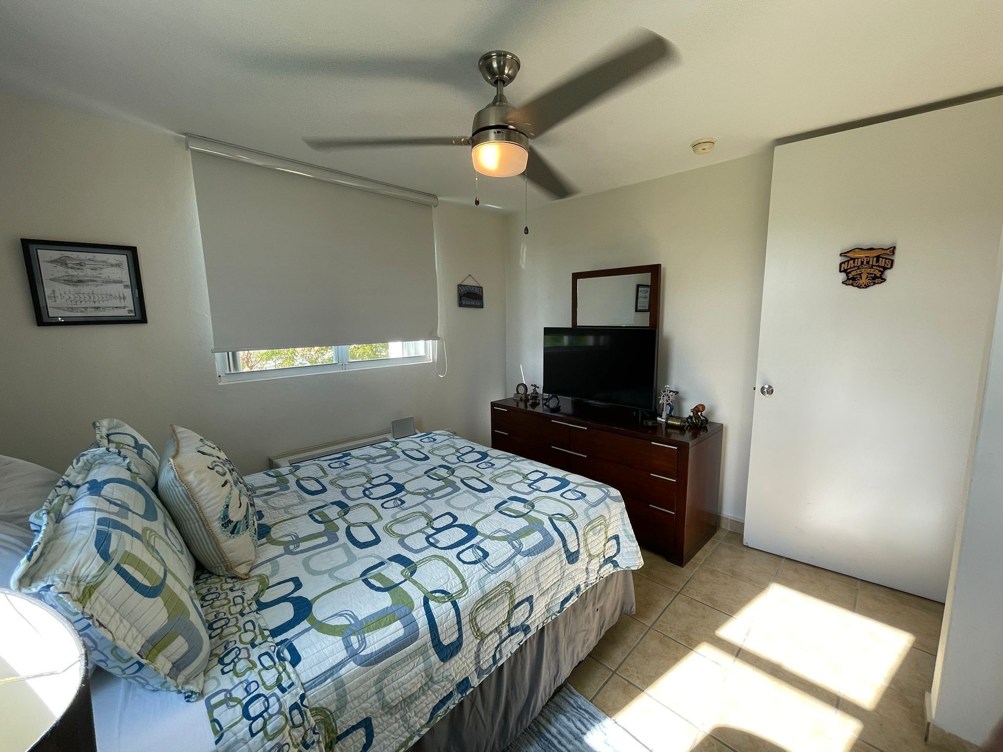 A bedroom with a bed, a dresser, a television, a ceiling fan, a window, and a door.
