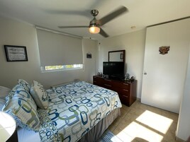 Un dormitorio con cama, cómoda, televisor, ventilador de techo, ventana y puerta.