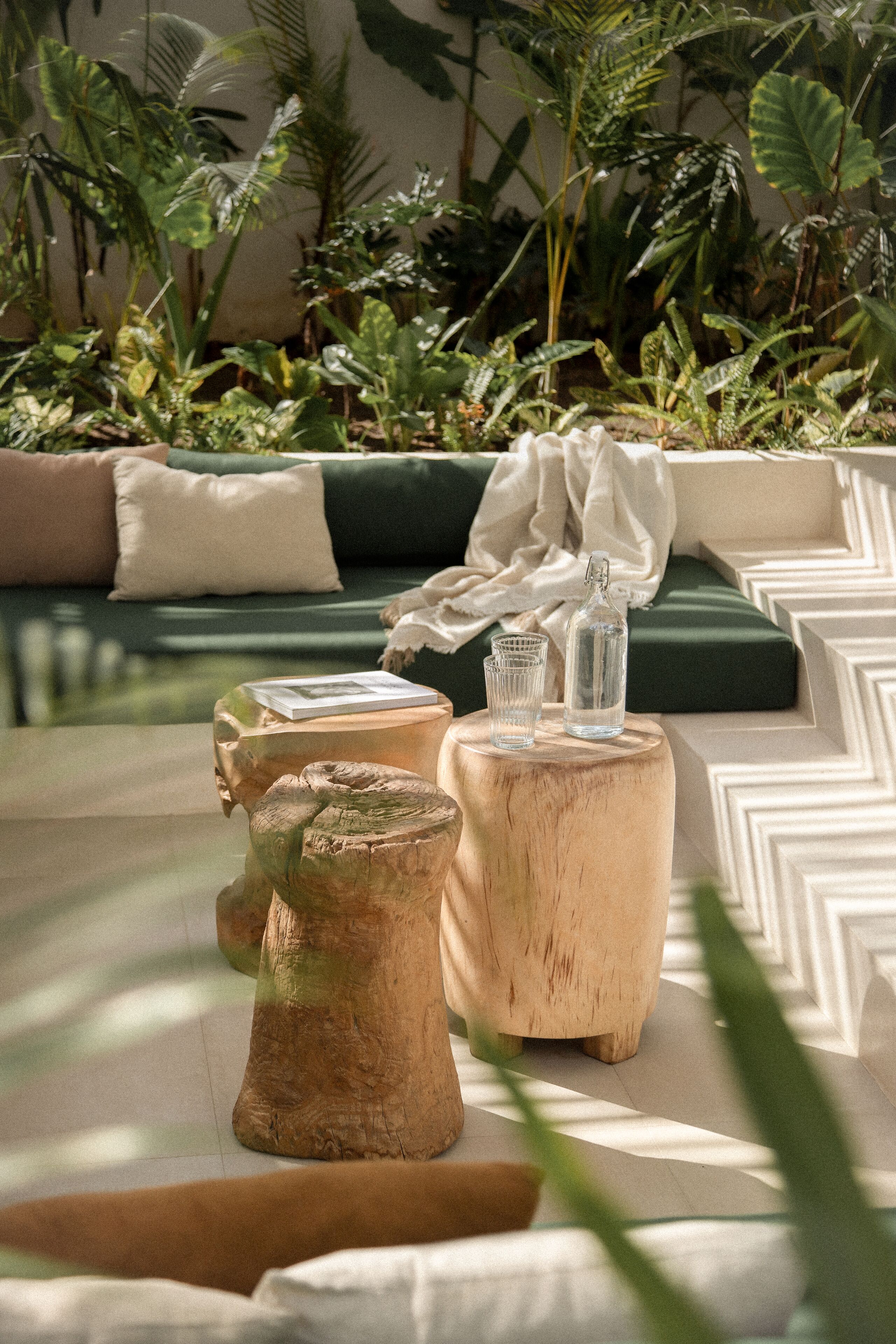 A wooden table with a bottle and glasses, a green sofa with cushions, and lush greenery in the background.