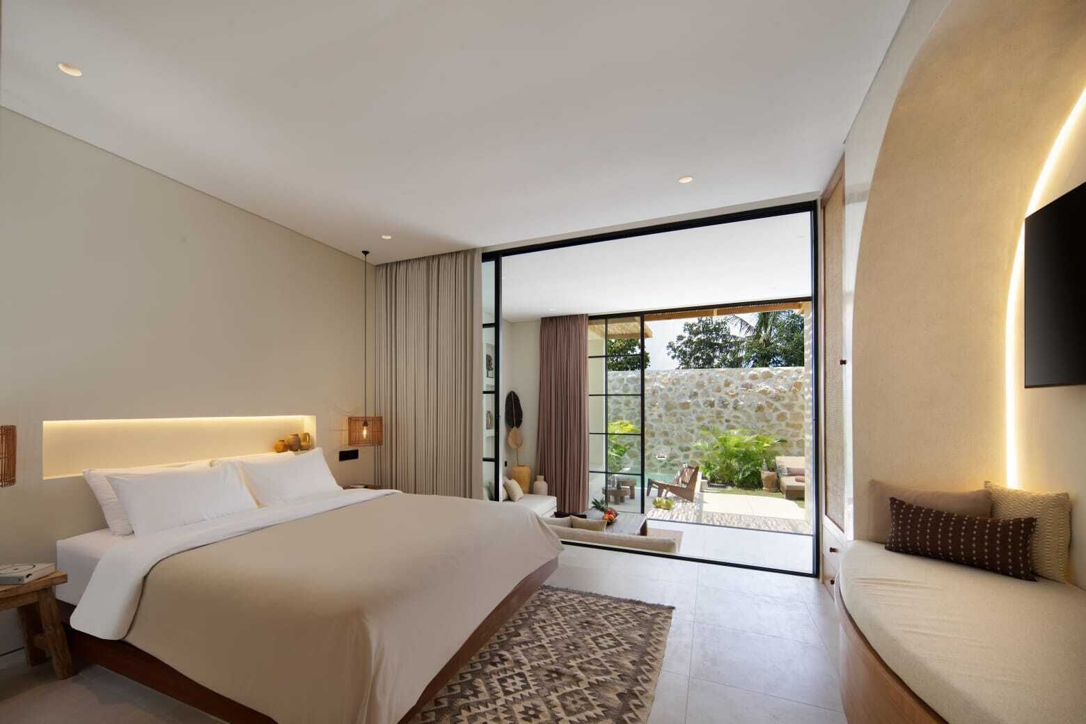 A modern bedroom with a large bed, a bedside table, a nightstand, a sofa, and a view of an outdoor patio.