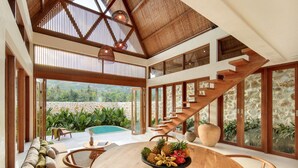 Un vaste espace intérieur doté d’un escalier en bois, d’une table à manger ornée de fruits et offrant une vue sur une piscine à l’extérieur.