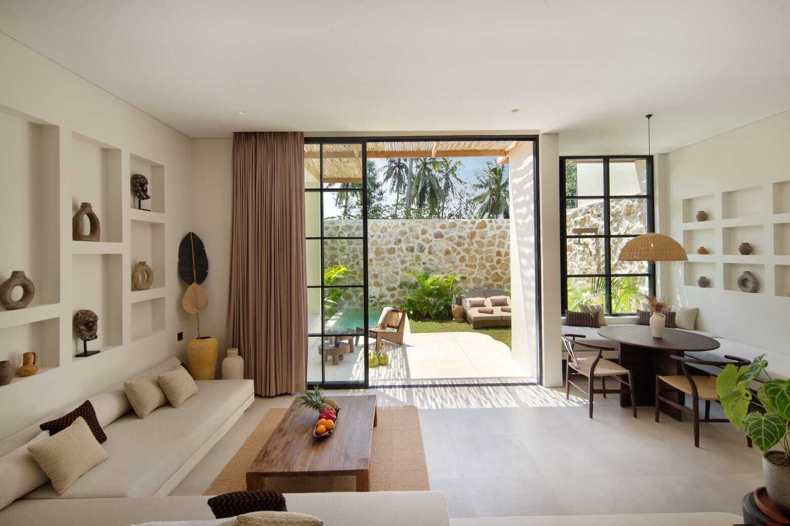 Una sala moderna con sofá, mesa de centro y comedor. Hay estanterías con adornos y vista al patio exterior.