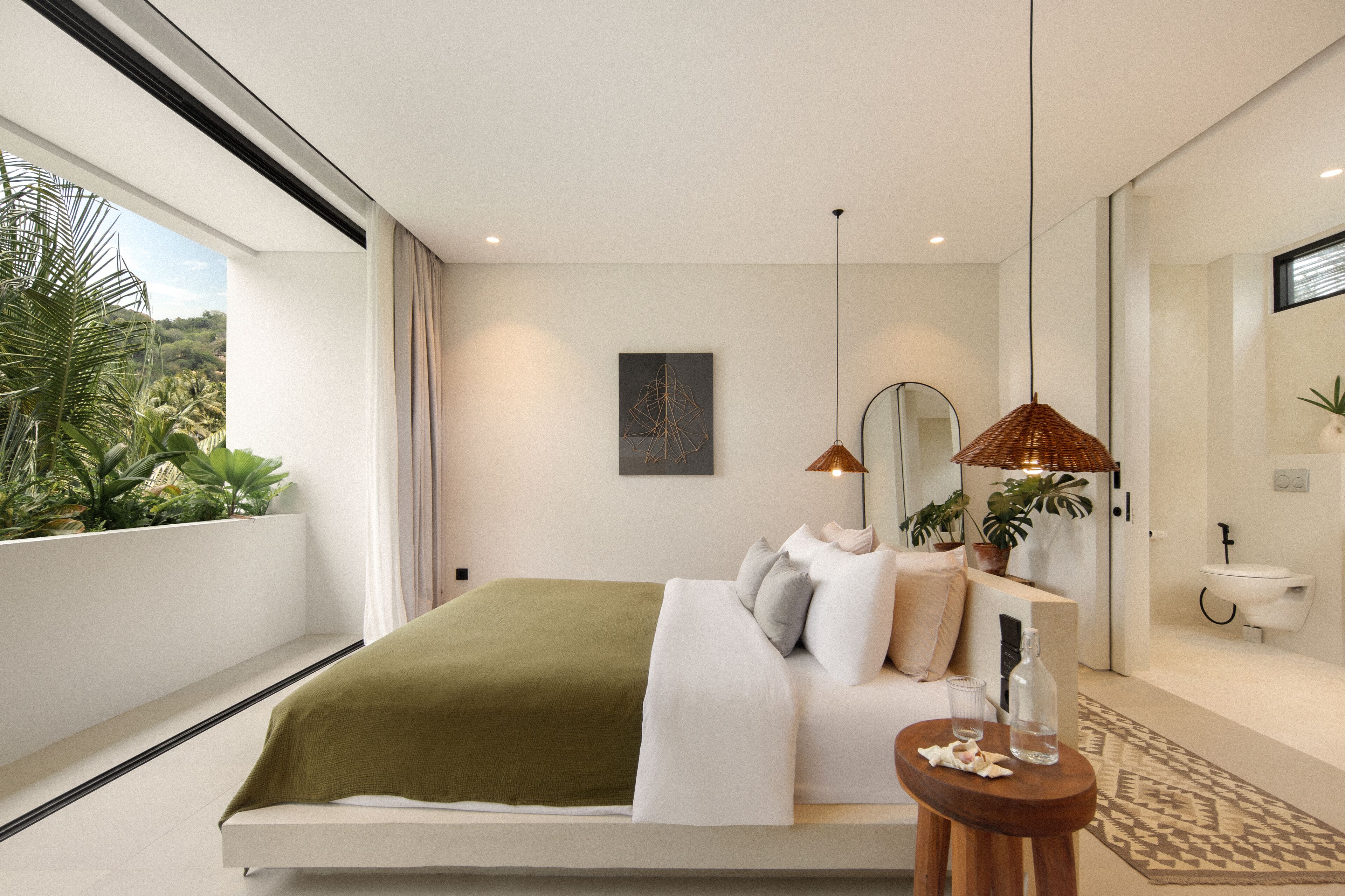 A modern bedroom with a large bed, a bedside table, a mirror, and a view of greenery outside.