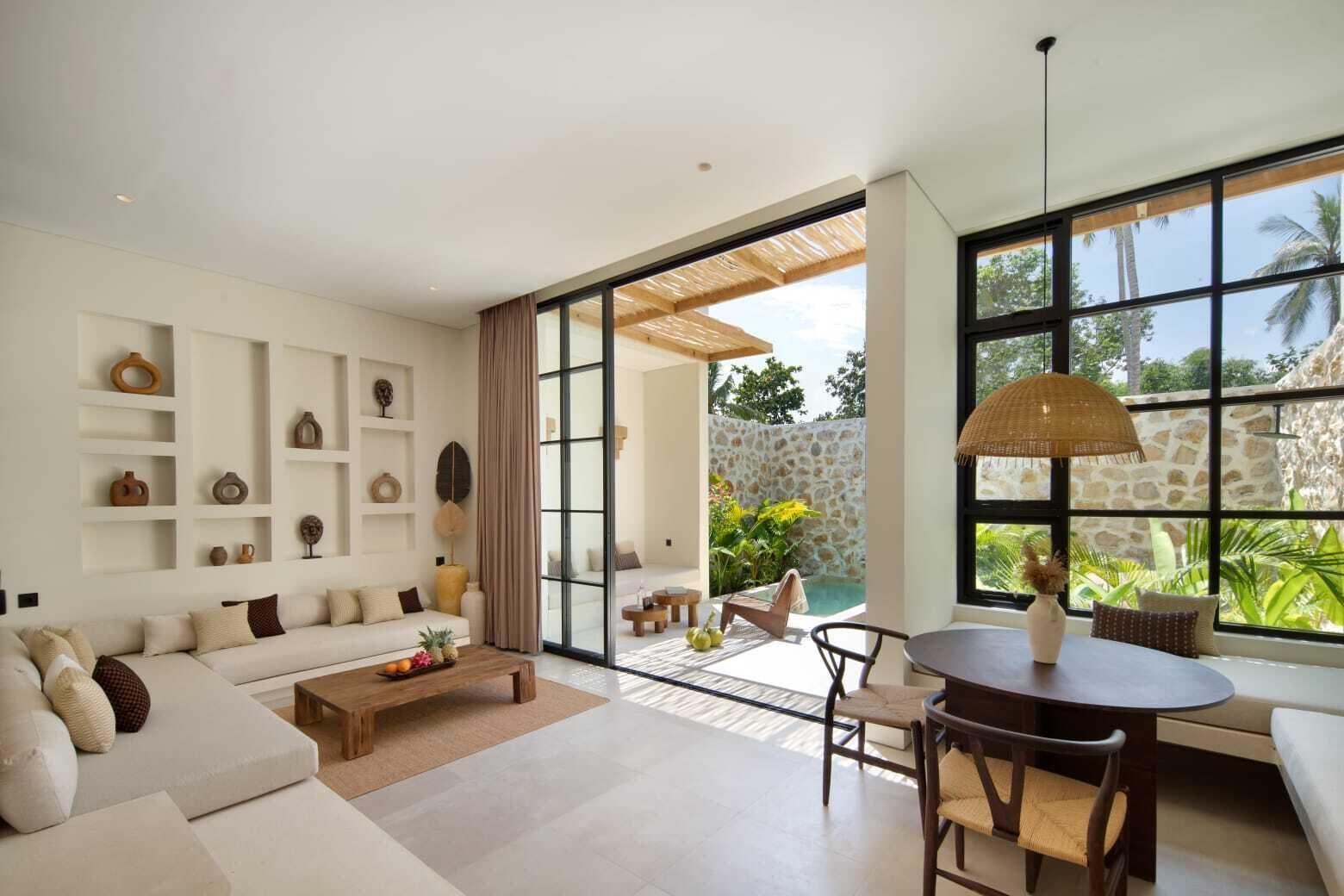 A modern living room with a sofa, coffee table, and dining area. There is a large window with a view of a stone wall and greenery.