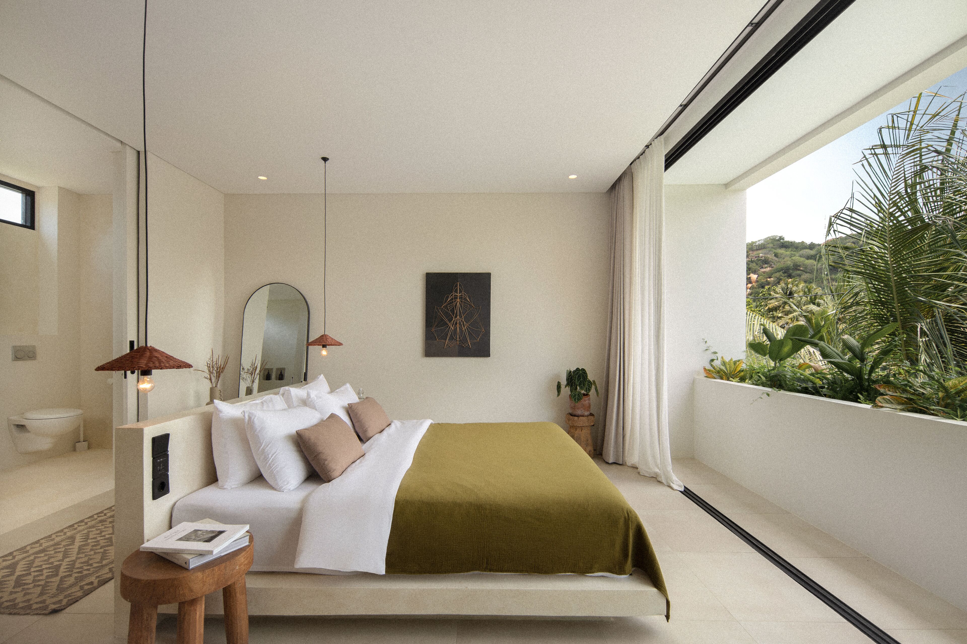 A modern bedroom with a large bed, a bedside table, a mirror, and a view of greenery outside.